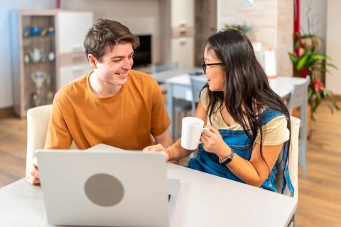 Young multiethnic couple smiling and looking at each other while using laptop and drinking coffee,