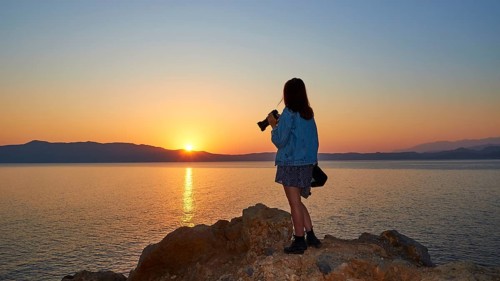 Sunrise, sun reflection on the sea, orange-blue cloudless sky, girl from behind with jeans jacket
