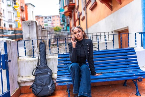 African american woman sitting on a city blue bench talking to the mobile with a guitar next to her