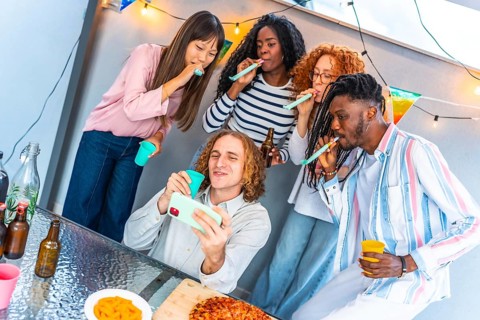Happy caucasian man taking selfie while friends blowing streamers during birthday rooftop party