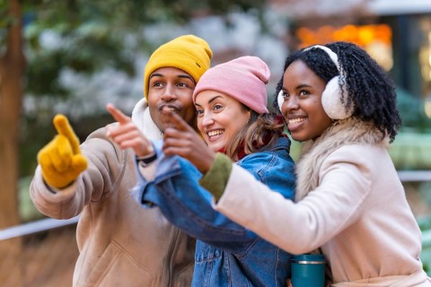 Three happy multi ethnic friends wearing winter clothes pointing their fingers at something