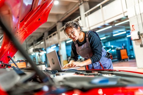 Young female mechanic working on a laptop while inspecting a car engine in a modern workshop,