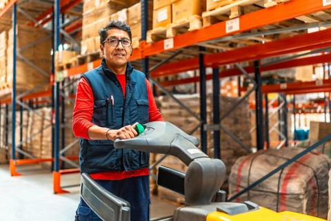Three quarter length photo of a happy latin man driving hand truck in a distribution warehouse