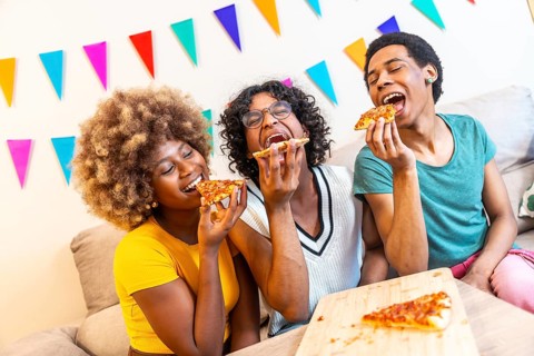 Multi-ethnic friends biting a slice of pizza having fun sitting on the sofa together at home