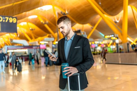 Businessman waiting for his flight, using smartphone and holding reusable coffee cup at the airport