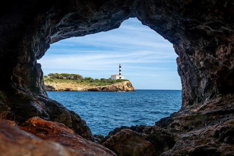 Portocolom lighthouse framed by the rocky opening of the arc natural cave, with blue sea and sky in