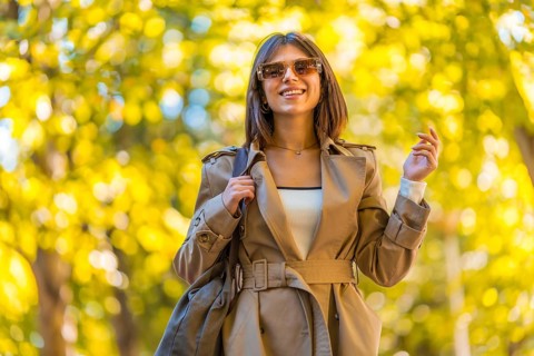 A woman wearing a tan coat and sunglasses is walking down a path. She is smiling and holding a