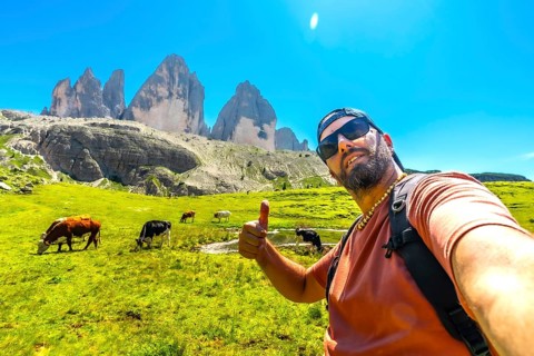 Hiker giving thumbs up while taking a selfie with cows grazing in a meadow with the tre cime di