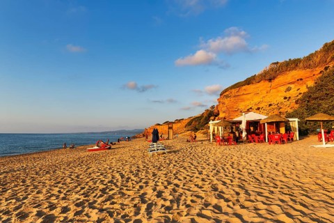 Beach café, red rocks illuminated by the evening sun, sandy beach with tourists, holiday by the