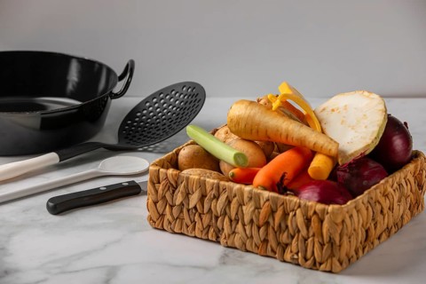 A woven basket full of vegetables such as soup vegetables, potatoes and onions next to kitchen