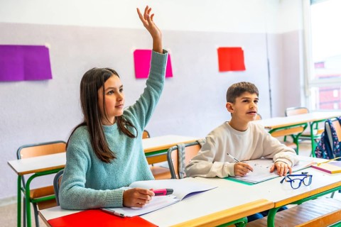 Girl raising hand to answer a question during class in elementary school while her classmate is