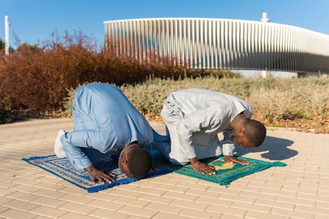 Two senegalese men wearing traditional dashiki clothing are kneeling on prayer rugs, performing