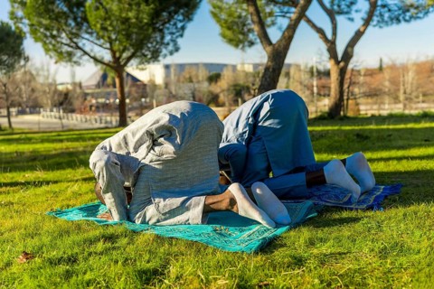 Two african muslim men are kneeling in prayer on prayer rugs in a park, wearing traditional