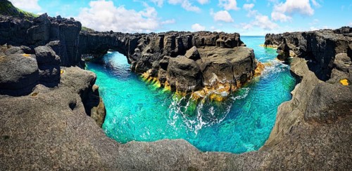 Panoramic view of rugged lava rock formations with a natural arch over turquoise sea under blue