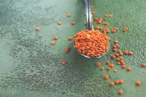A spoon holds vibrant red lentils, with more lentils scattered around on a textured green surface.