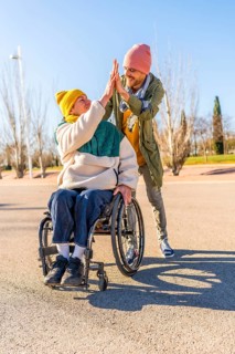 Two friends celebrating something with a high five in a park, one of them using a wheelchair,