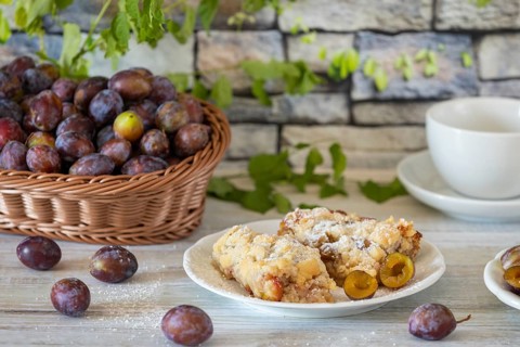 Plum cake on a plate next to a basket with fresh plums and a cup in the background, on a white