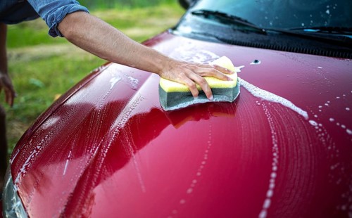 Close-up of a hand washing the hood of a red car with a sponge and soap