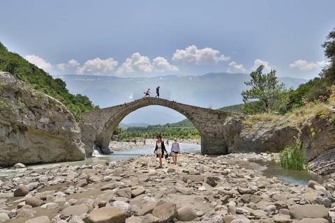 Two young woman walking barefoot in the riverbed over large stones, Ottoman stone bridge, Kadiut