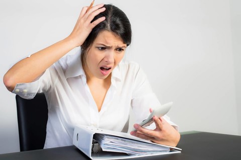 Businesswoman sitting at her desk with a large office binder looking at her calculator in horror or