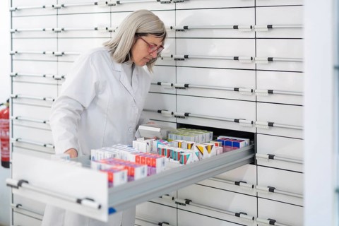 A Caucasian female pharmacist with glasses dispenses ordered medications from the dispensary,