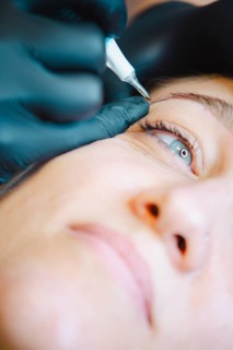 Beautician applies permanent makeup to a woman's eyebrow in a busy salon, showcasing precision and