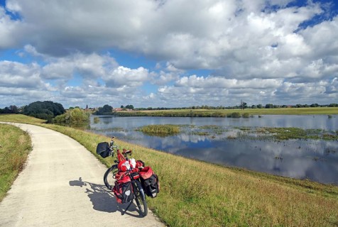 A bicycle with panniers stands on a path next to a calm river and under a slightly cloudy sky,