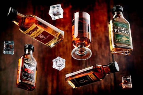 Four whisky bottles and a glass of whisky with floating ice cube cubes against a wooden background