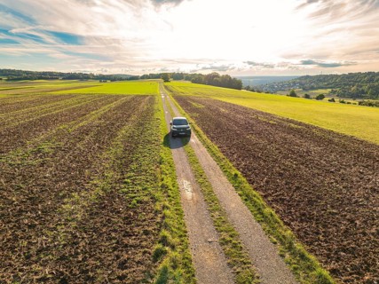 Black car driving on a long, straight country lane through scenic fields under a cloudy sky, car
