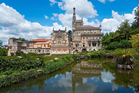 Historisches Schloss mit reflektiertem Bild im See, umgeben von Bäumen und bewölktem Himmel,
