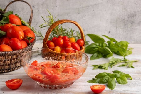 Glass bowl with prepared tomato sauce and sliced tomatoes, surrounded by fresh tomatoes in baskets,