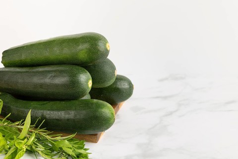 Fresh courgettes stacked on a wooden board with fresh herbs against a light-coloured background