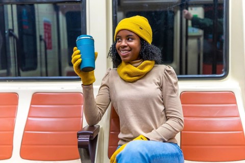 Smiling young woman wearing yellow winter accessories holding reusable mug while commuting on