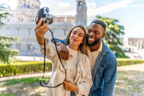 Happy multi-ethnic couple taking selfie with digital camera in a park with views on downtown