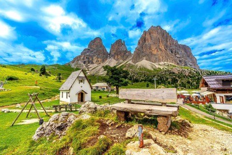 Picturesque landscape featuring a wooden bench, a small chapel, and the majestic peaks of the sella