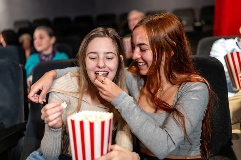 Two female friends enjoying a movie night, sharing popcorn and laughter in a crowded cinema