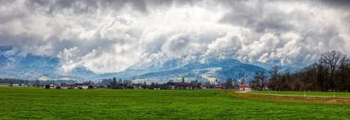 Green meadow and small village in the foreground, dramatic clouds, mountains in the background, Bad