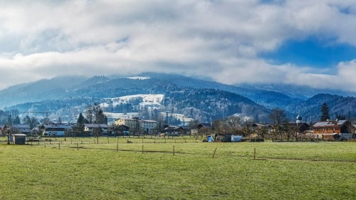 A village in front of snow-covered mountains under a cloudy sky on a green meadow, Bad Feilnbach,