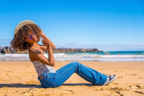 Beauty latin woman sitting on the sand of a beach playing with sun hat and smiling at camera