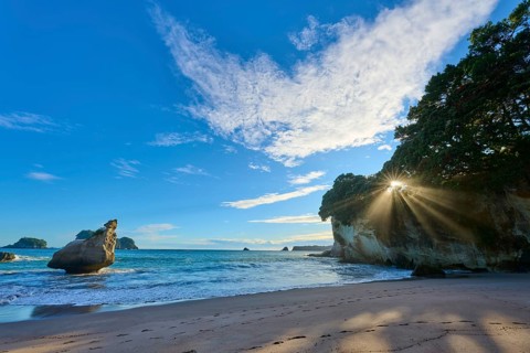 Landscape with sandy beach and bright sun behind rocks by the sea, Cathedral Cove, Coromandel,