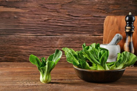 A bunch of vibrant bok choy sits in a bowl alongside a single piece on a wooden table, complemented