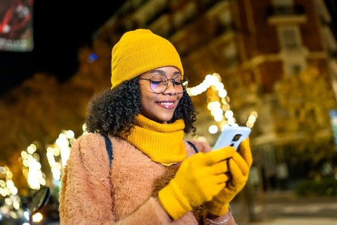 Smiling young woman wearing yellow winter clothes using smartphone in a city street at night with