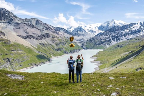 Two friends throwing yellow hats in the air, mountaineers in front of a blue mountain lake, Lac des