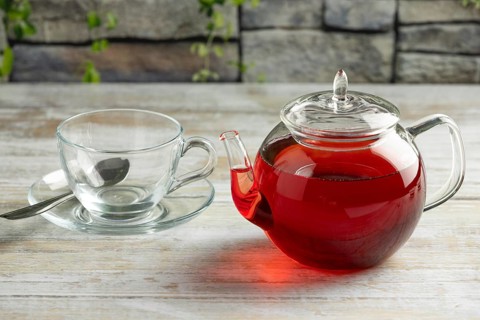 A teapot with red tea next to an empty glass cup with spoon on a table in front of a stone