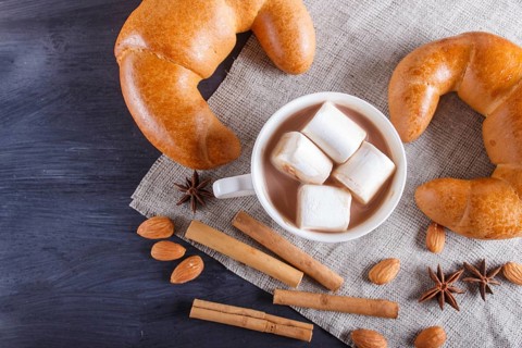 A cup of hot chocolate with marshmallow, croissants and spices on linen napkin and black wooden