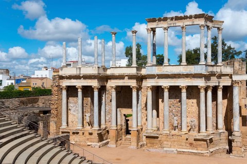 Ancient Roman theatre with columns and stairs, in the background a modern city under a blue sky,