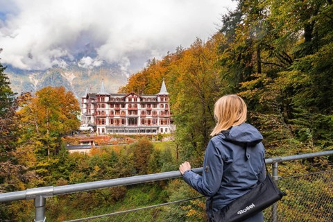 A woman looks from a railing at a large hotel surrounded by autumnal forest, Lake Brienz, Giessbach