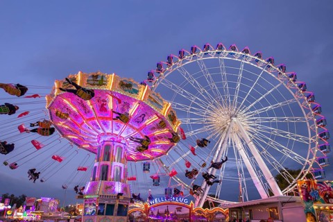 A lively funfair at dusk, illuminated by a large Ferris wheel and a colourful chain carousel,