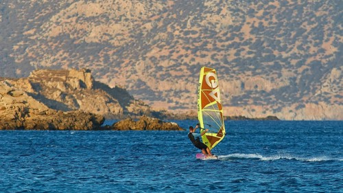Person windsurfing in front of rocky coastal scenery and blue mountains in the background,