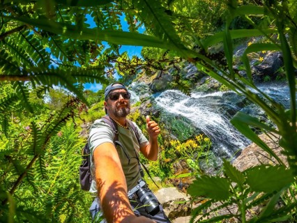 A man is taking a picture of himself in a forest. He is wearing a hat and a backpack